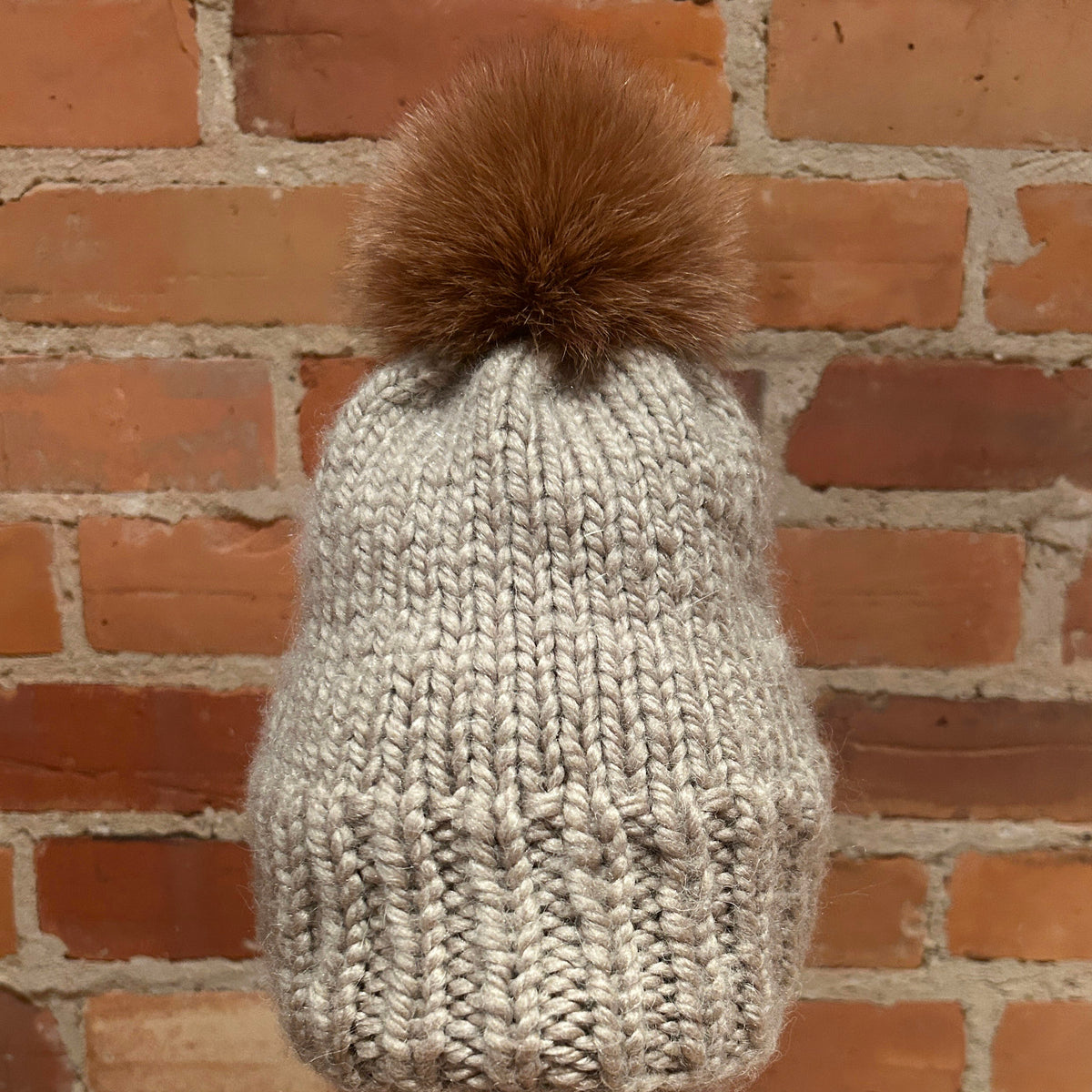 A chunky knit beanie hat in light oatmeal beige with a large brown fox-fur pom-pom, displayed on a mannequin head against a red brick wall background.