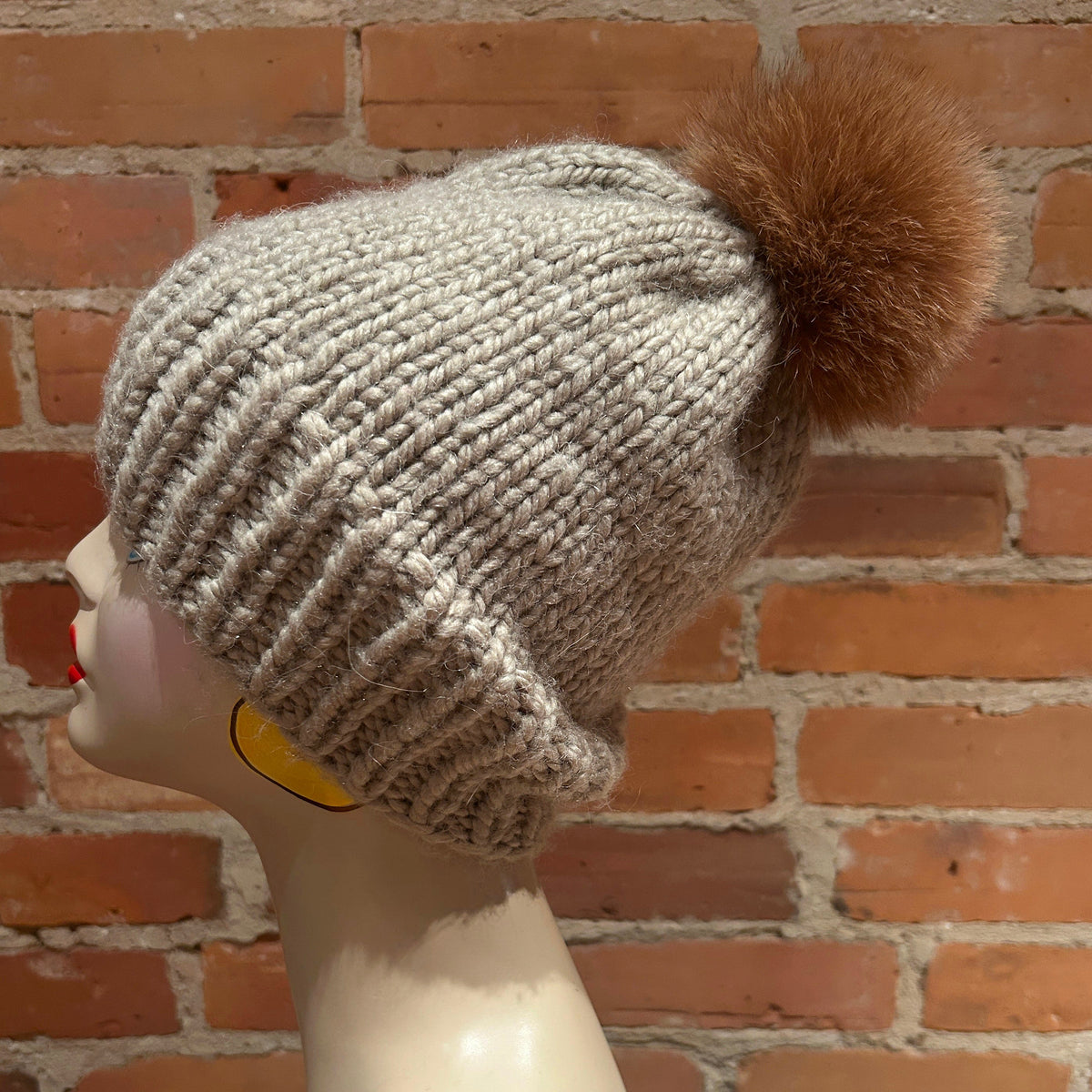 A chunky knit winter beanie in light oatmeal gray with a large brown fox-fur pom-pom, displayed on a mannequin head against a red-brown brick wall background.