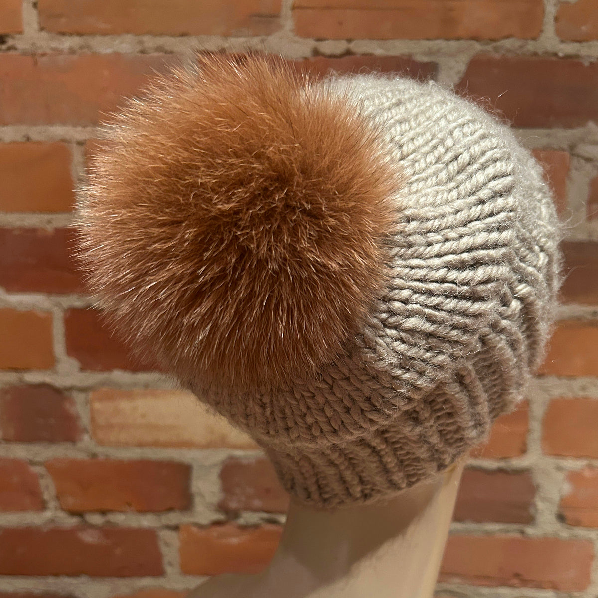 A close-up of a knit beanie hat on a mannequin head, featuring a chunky ribbed light beige body and a large fluffy camel-brown fox fur pom-pom, set against a rustic red brick wall background.