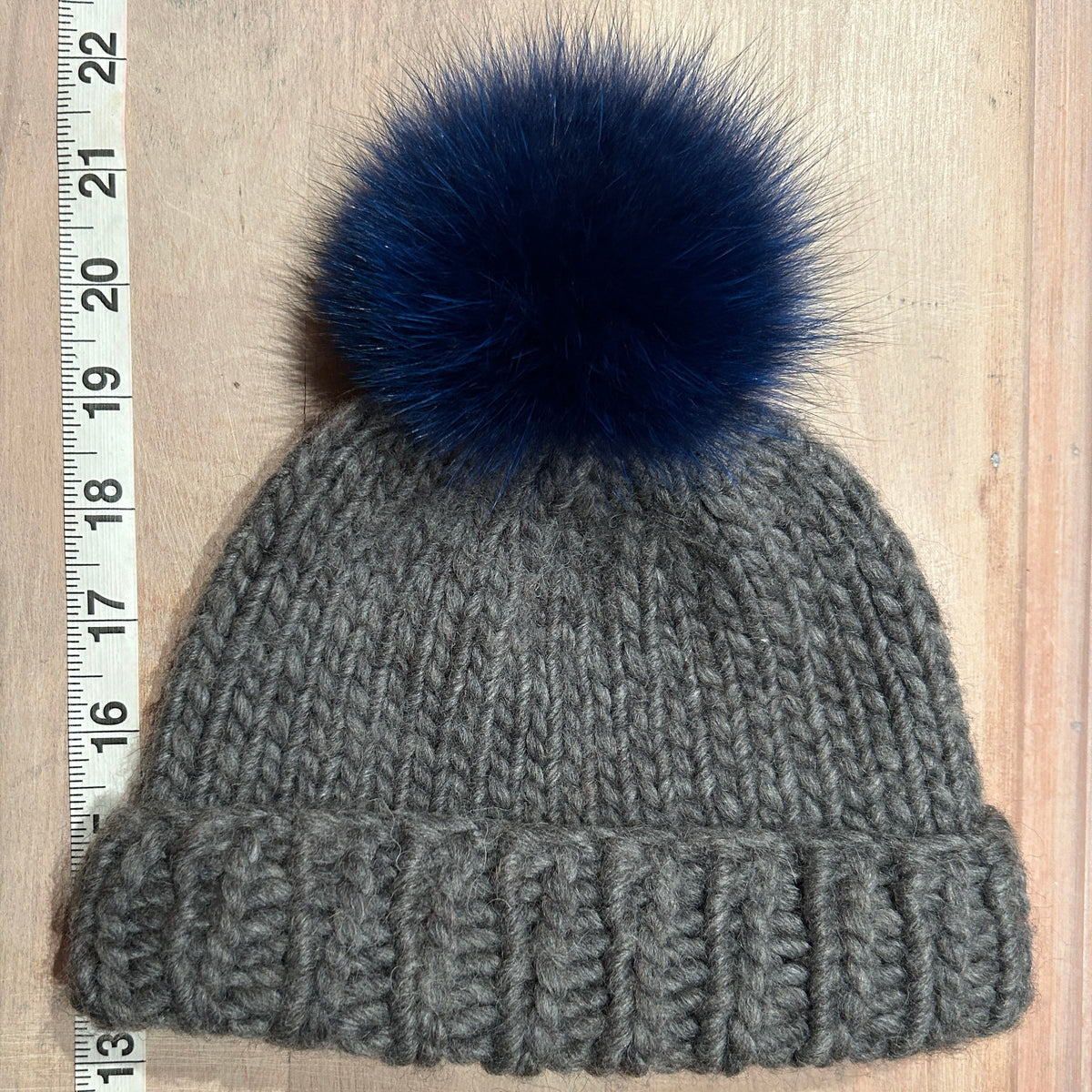 A small gray ribbed knit beanie with a small dark navy blue fox fur pom-pom, shown flat on a light wood background next to a measuring ruler.