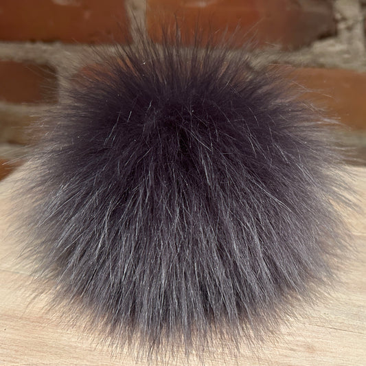 A fluffy dark gray faux fur pom-pom ball resting on a light wood surface with a blurred brick wall background.