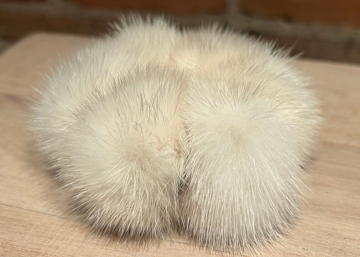 A pair of fluffy mink scrunchie, in a cream/off-white color, resting on a light wood surface.