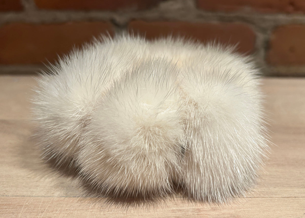 A pair of fluffy mink fur scrunchie  in a cream-white color, resting on a light wood surface with a brick wall background.