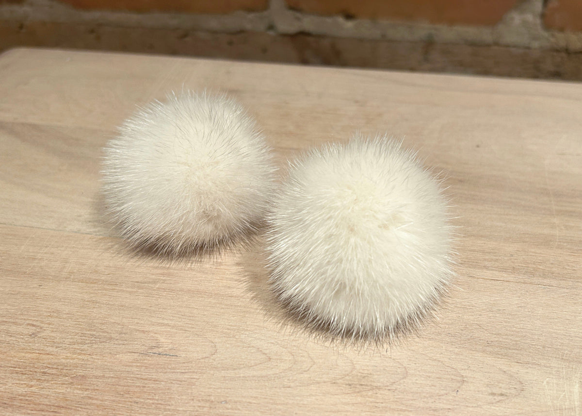 Two fluffy cream mink fur pom-poms resting on a light wood tabletop with a brick wall background.