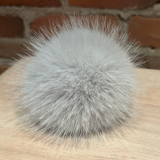 A fluffy light gray faux fur pom-pom ball resting on a light wood surface with a blurred brick wall background.