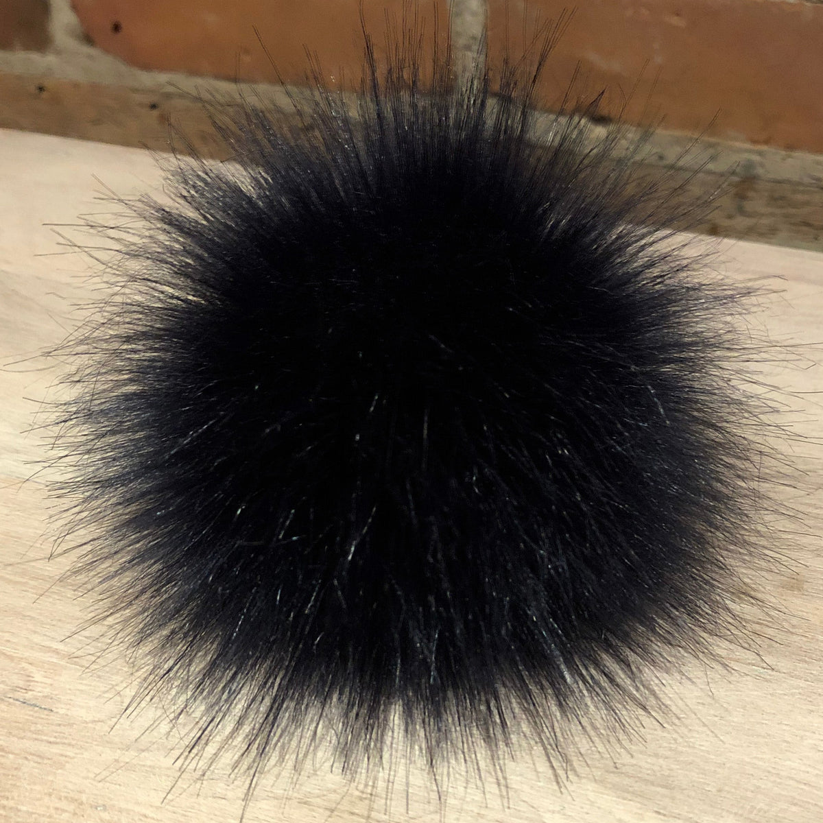 A round, fluffy navy blue faux fur pom-pom accessory resting on a light-colored wooden surface with a brick wall background.