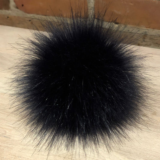 A round, fluffy navy blue faux fur pom-pom accessory resting on a light-colored wooden surface with a brick wall background.