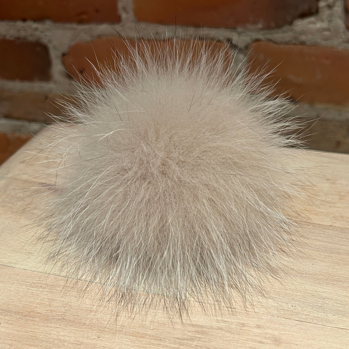 A fluffy fox fur pom-pom in a light beige color, resting on a light wood surface with a brick wall background.