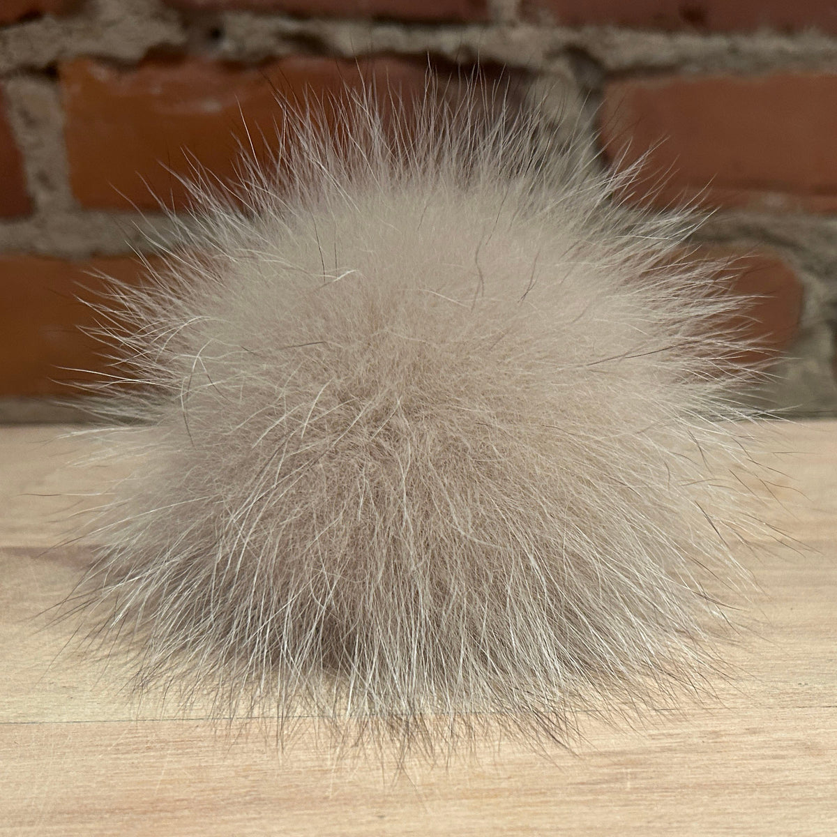A fluffy fox -fur pom-pom ball in a light taupe-beige color, resting on a light wood surface with a blurred brick wall background.