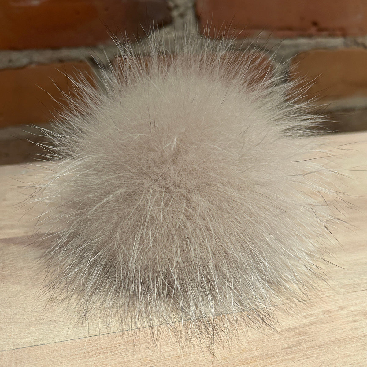 A fluffy fox-fur pom-pom ball in a light taupe-beige color, resting on a light wood surface with a blurred brick wall background.