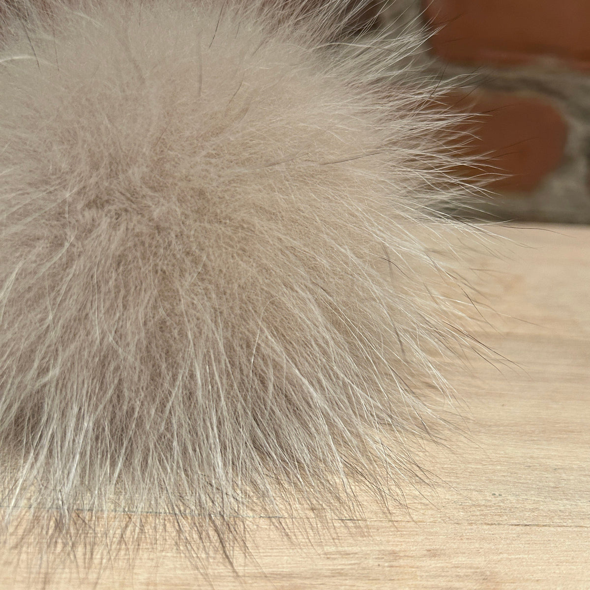 A close-up of a fluffy fox fur pom-pom accessory in a light taupe-beige color, resting on a light wood surface with a blurred brick wall background.