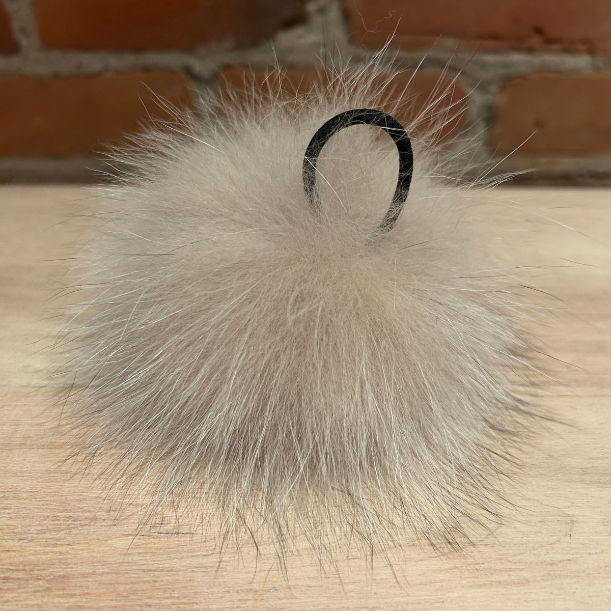 A fluffy fox -fur pom-pom keychain or bag charm in a light beige-gray color, featuring a black plastic hanging loop, resting on a light wood surface with a blurred brick wall background.