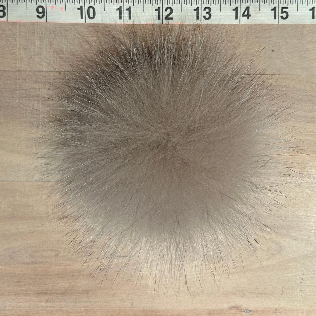 A round fluffy fox fur pom-pom in a light taupe-brown color, shown on a light wood surface with a measuring ruler at the top for scale.