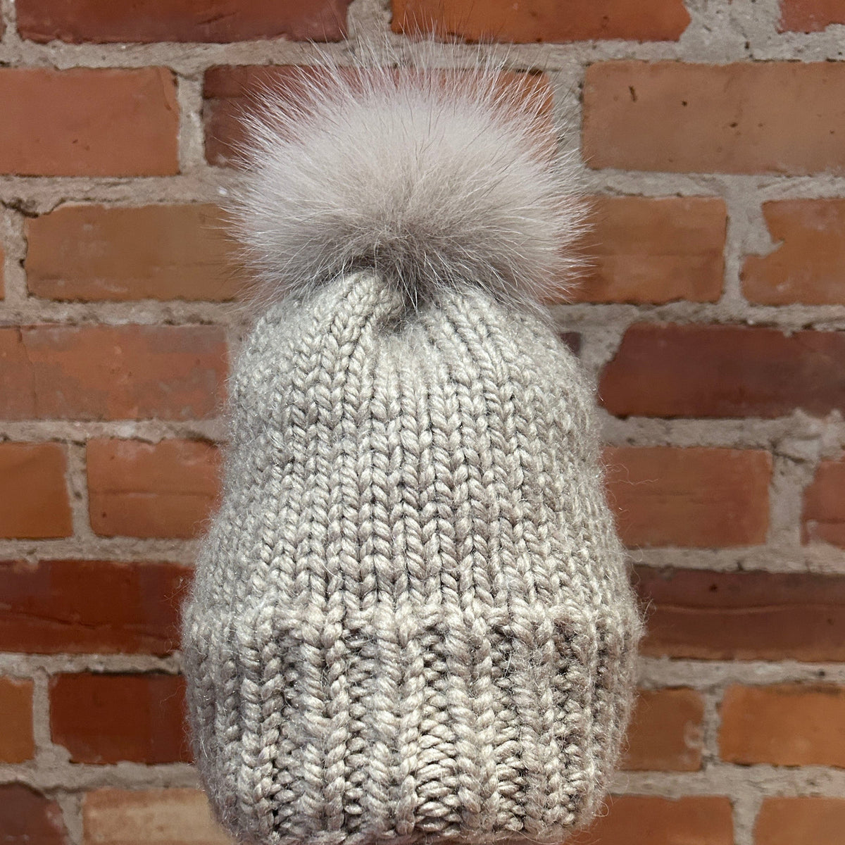 A light gray knit beanie with a fluffy light gray pom-pom, displayed on a mannequin head with red lipstick in front of a red brick wall.
