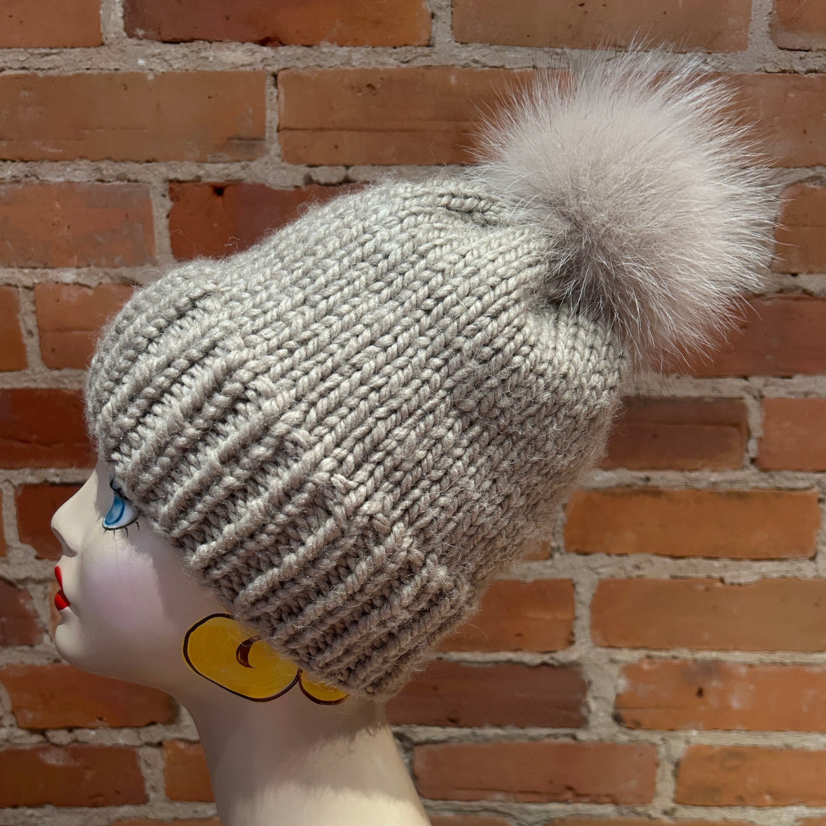 A light gray knit beanie with a large fluffy pom-pom, displayed on a mannequin head against a red brick wall background.