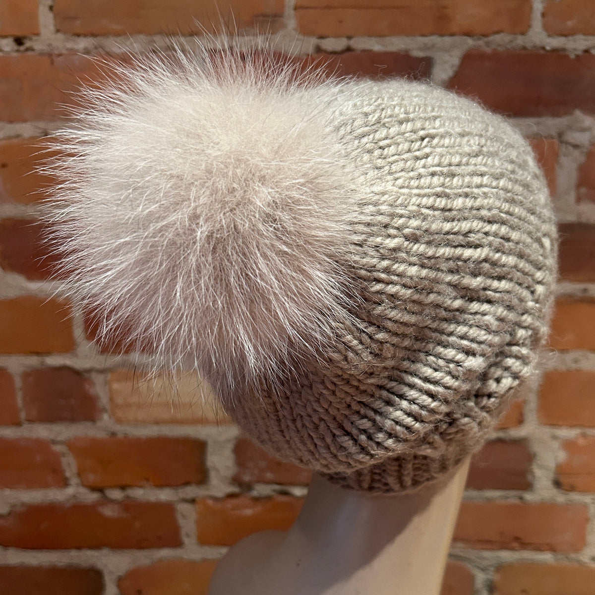 A chunky knit beanie hat in a light oatmeal gray color with a large fluffy fox-fur pom-pom, displayed on a mannequin head against a brick wall background.