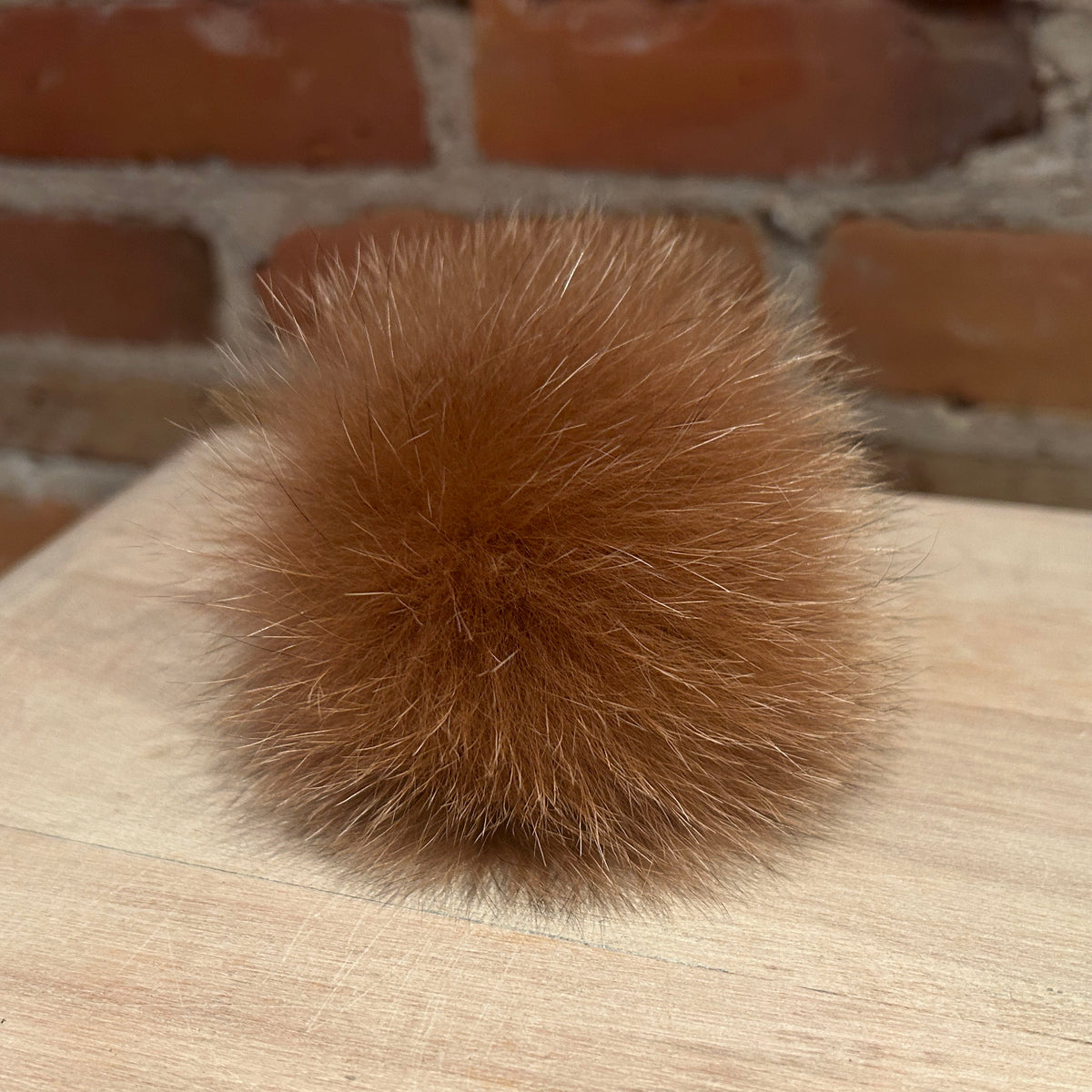 A fluffy ginger fox fur pom-pom ball resting on a light wood surface with a blurred brick wall background.