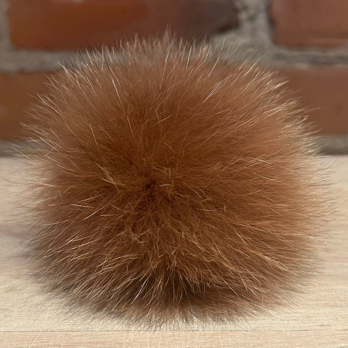 A fluffy, round ginger fox-fur pom-pom ball resting on a light wood surface with a blurred brick wall background.