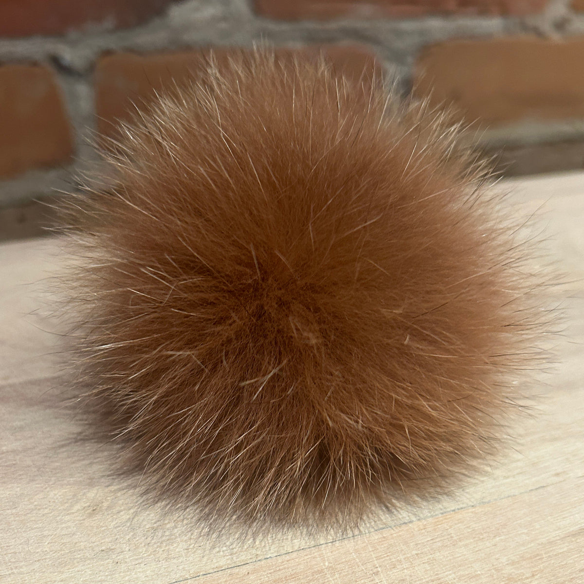 A fluffy ginger fox fur pom-pom ball resting on a light wood surface with a blurred brick wall background.