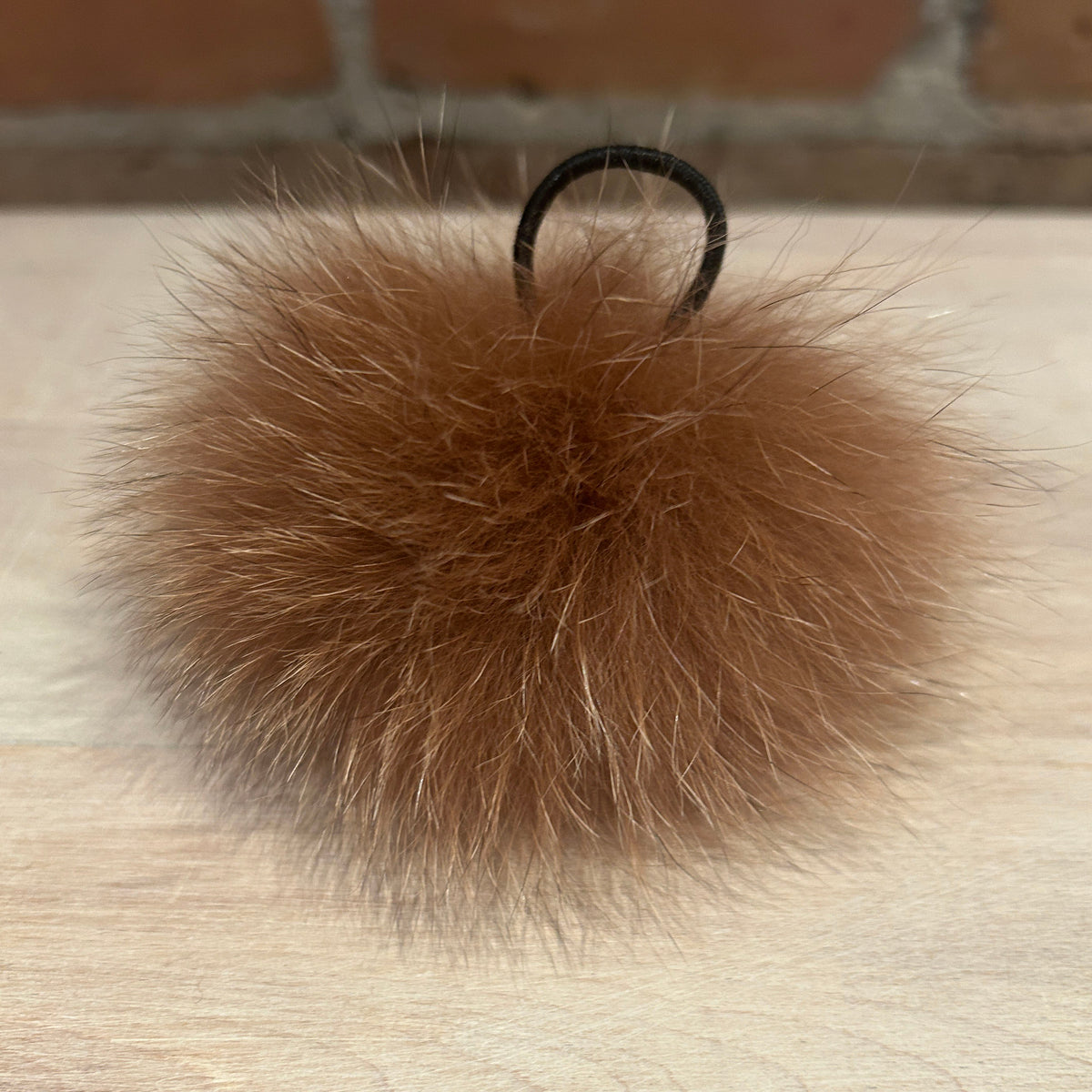A fluffy ginger fox-fur pom-pom keychain or bag charm with a black hanging loop, resting on a light wood surface with a brick wall in the background.