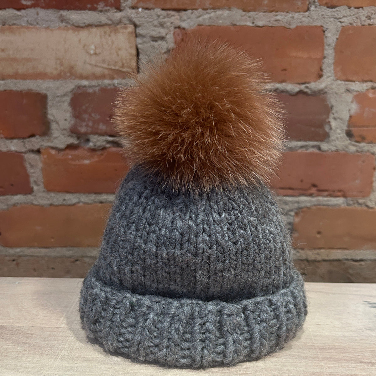 A small gray knit beanie hat with a small brown ginger fox fur pom-pom, displayed on a light wooden surface against a red brick wall background.