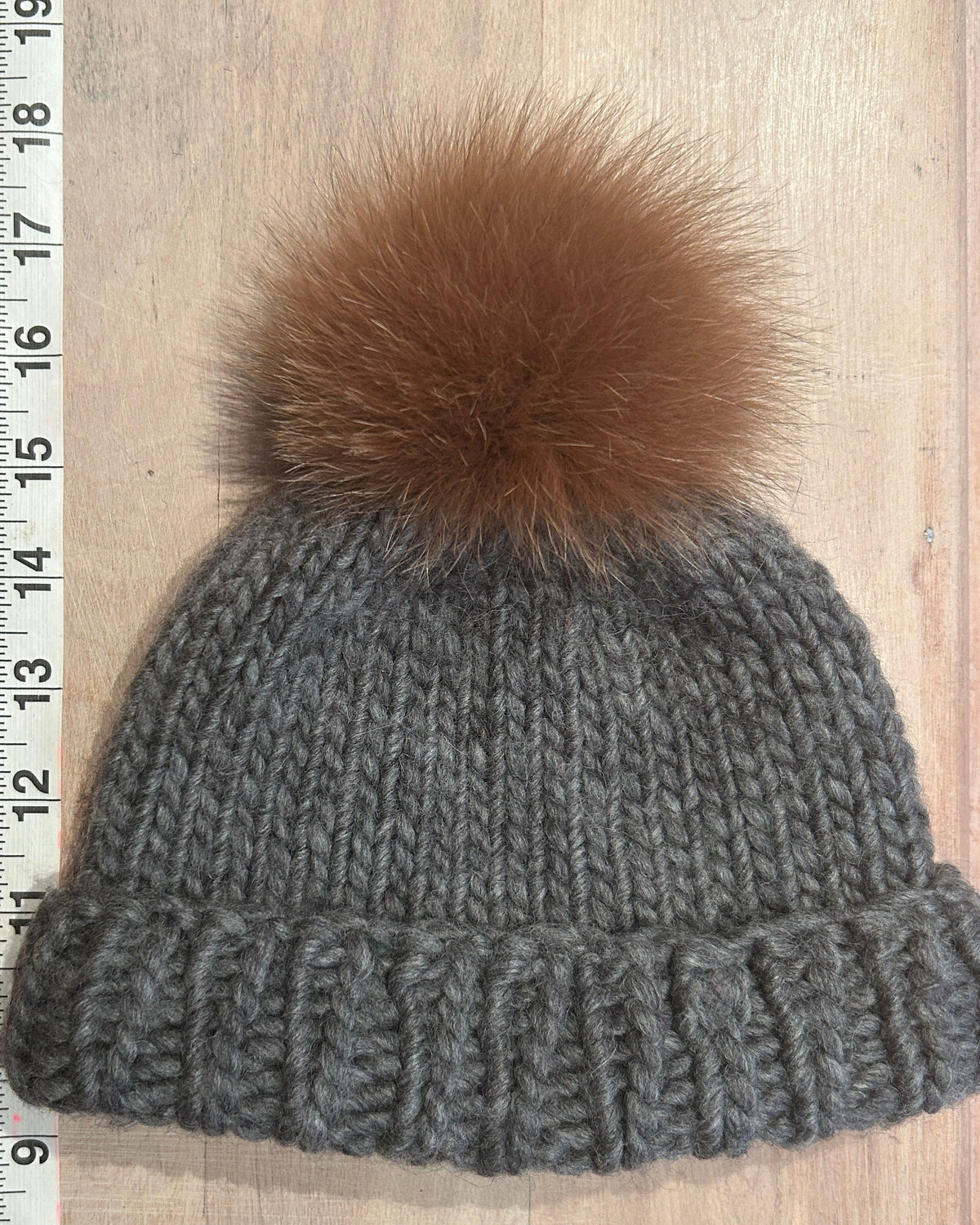 A small gray knit beanie with a small ginger fox-fur pom-pom, laid flat on a light wood surface next to a measuring ruler.