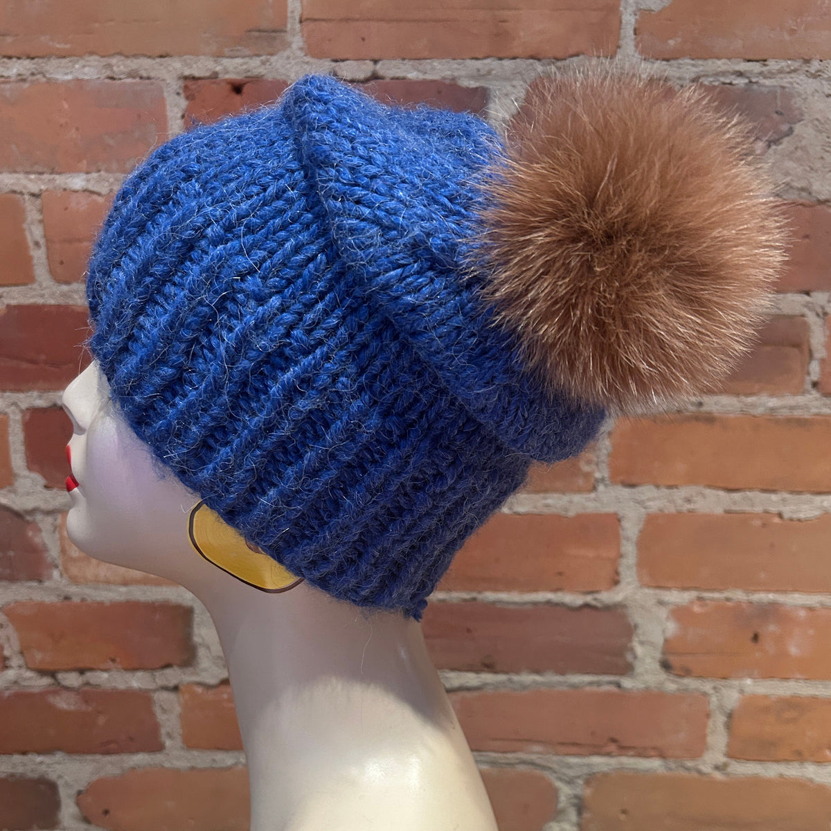 A blue ribbed knit beanie hat with a small ginger fox fur pom-pom, displayed on a mannequin head against a red brick wall background.