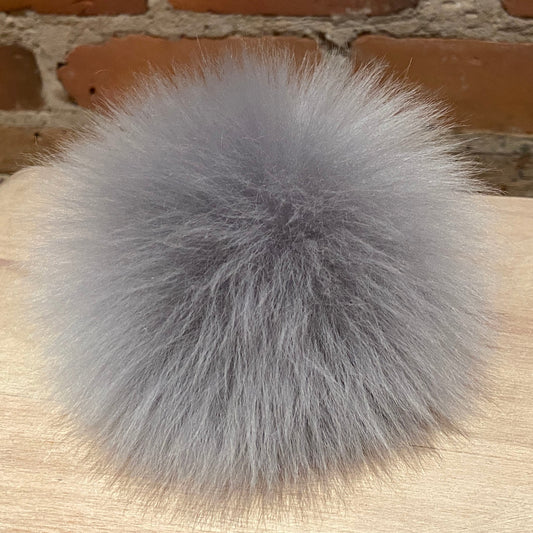 A fluffy gray lamb fur pom-pom accessory resting on a light wood surface with a brick wall background.