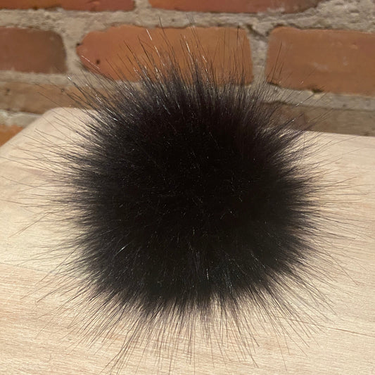 A black fur pom-pom accessory resting on a light wood surface with a brick wall background.