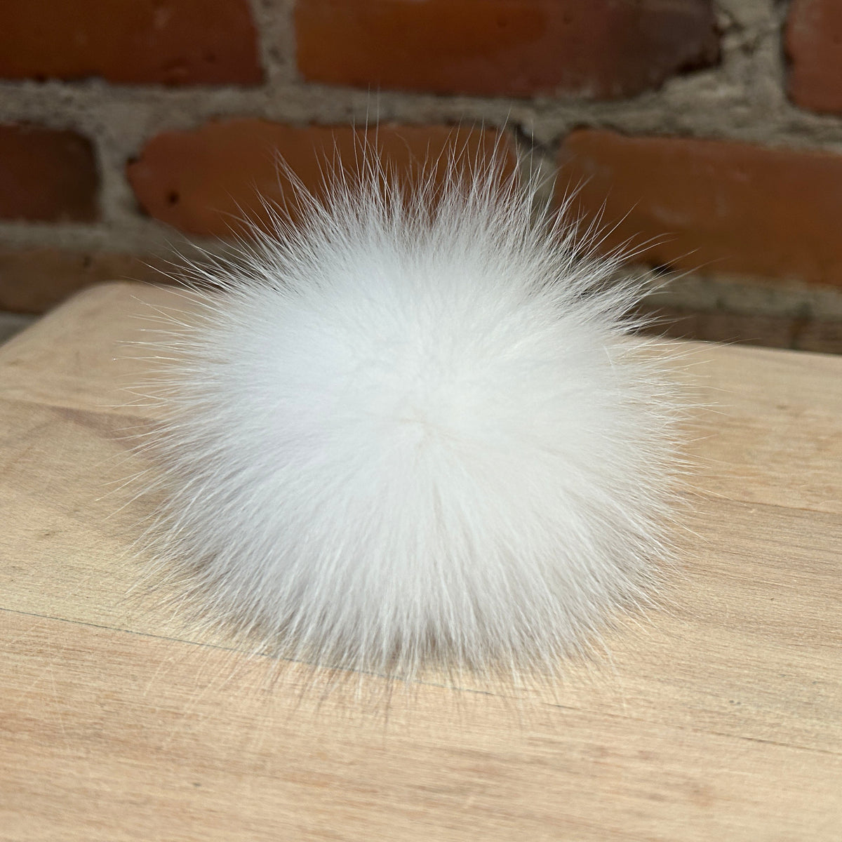 A fluffy white fox fur pom-pom ball resting on a light wood surface with a brick wall background.
