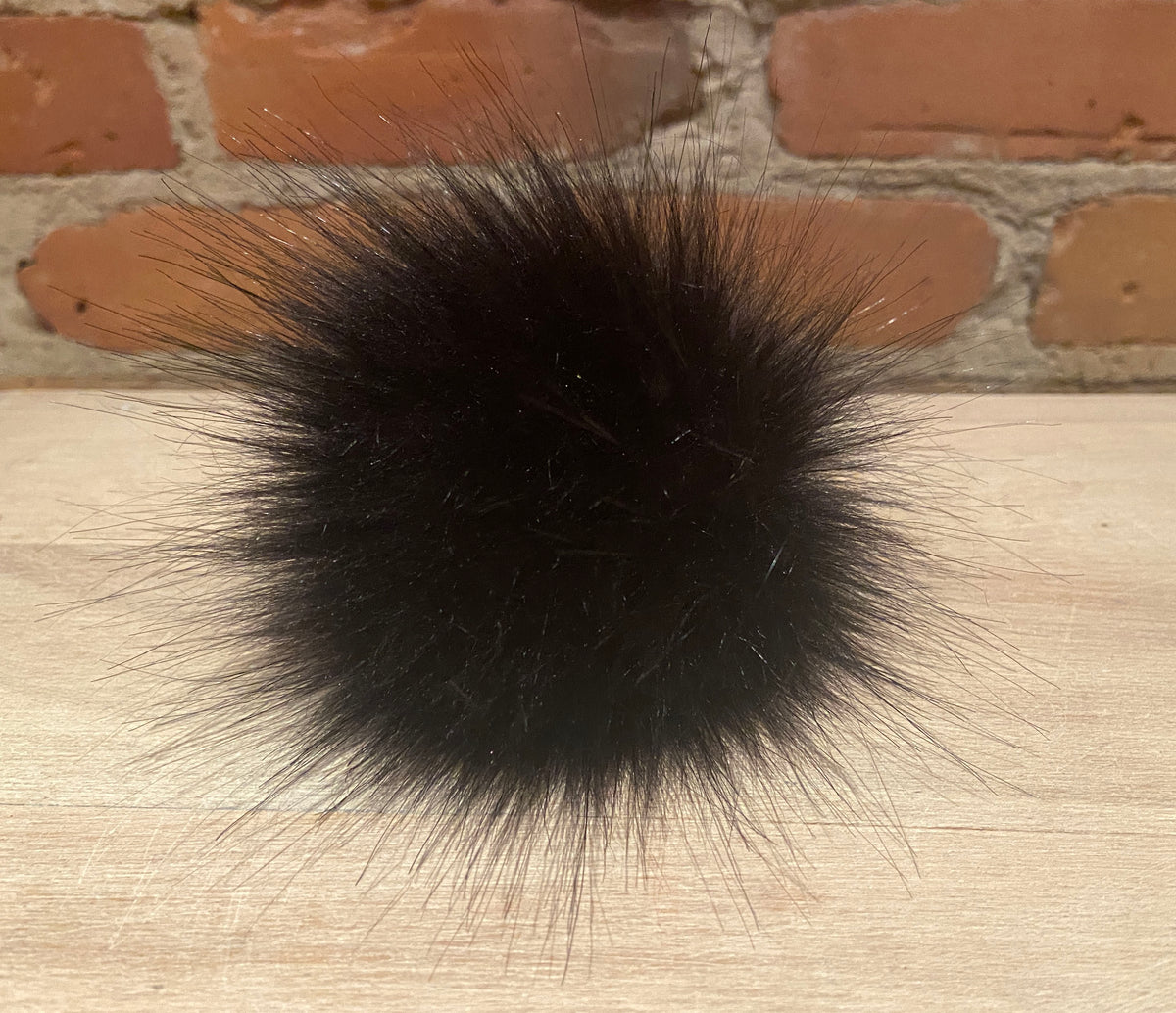 A black faux fur pom-pom ball accessory resting on a light wood surface with a brick wall background.