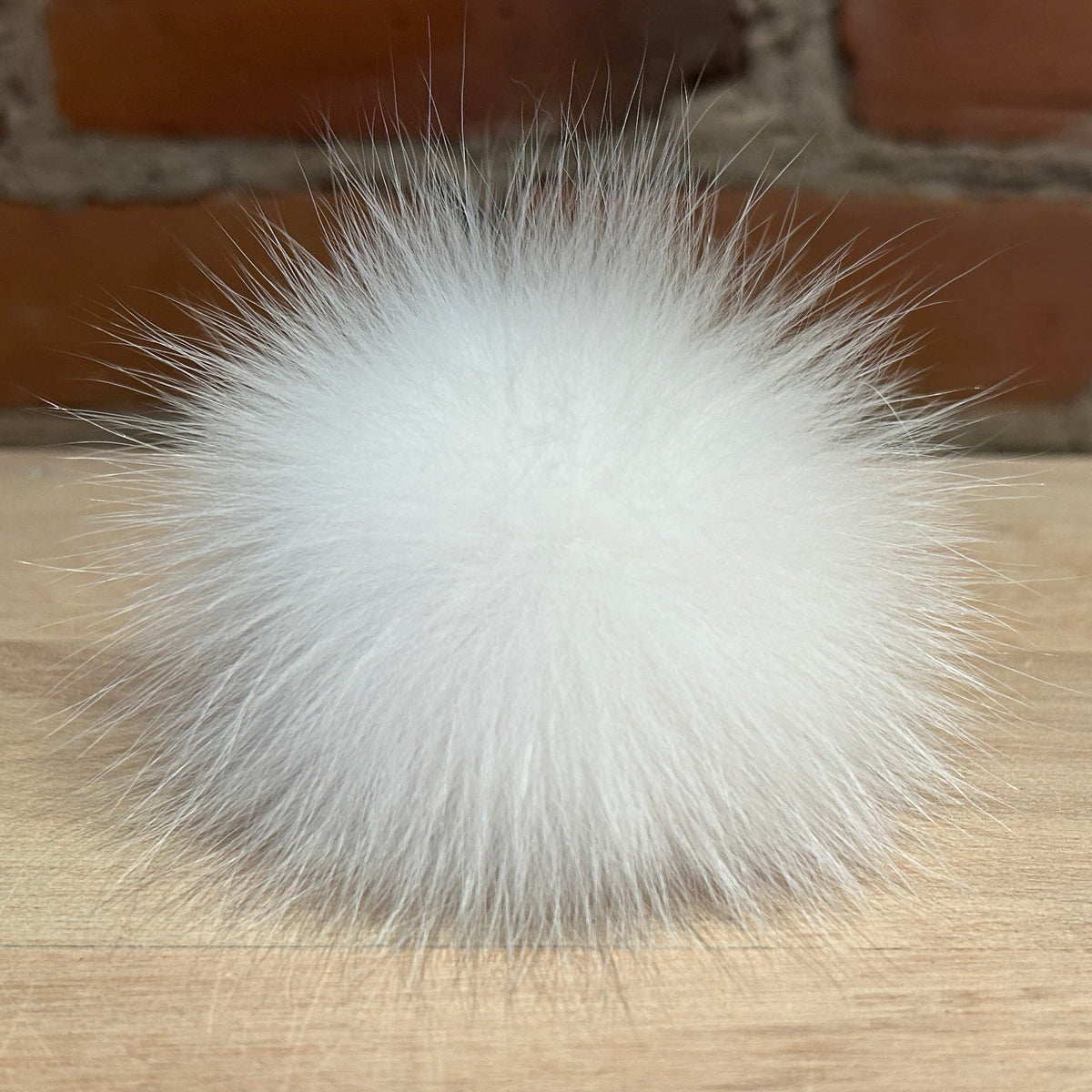 A fluffy white fox fur pom-pom ball resting on a light wood surface with a blurred brick wall background.