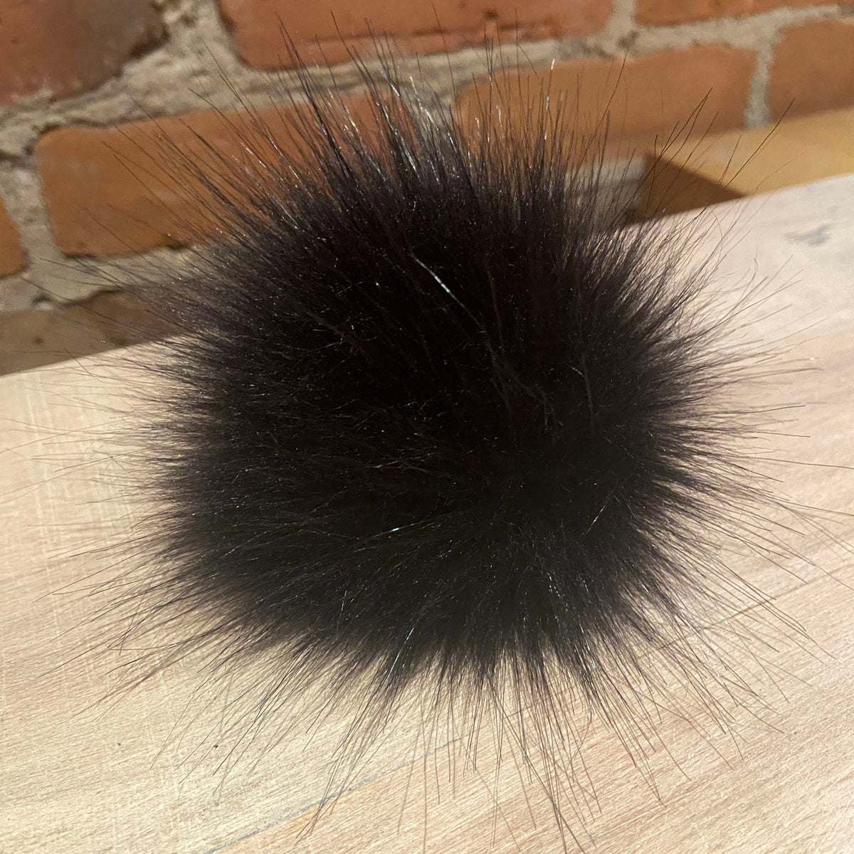 A round, fluffy black faux fur pom-pom resting on a light-colored wooden surface with a brick wall in the background.