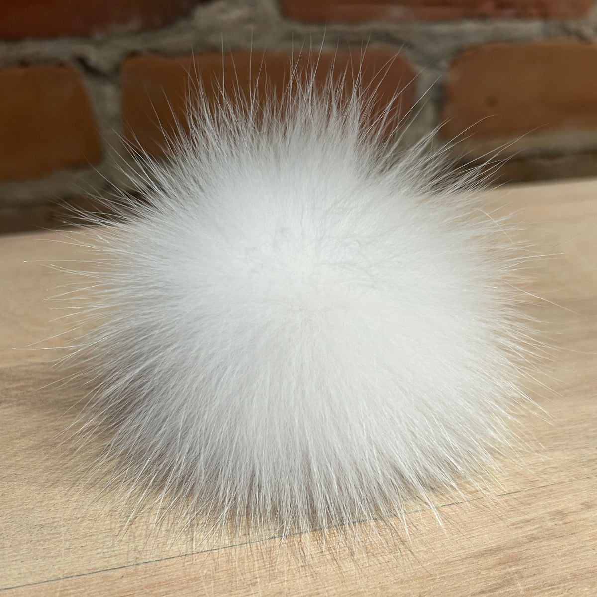 A fluffy white fox fur pom-pom ball resting on a light wood surface with a blurred brick wall background.