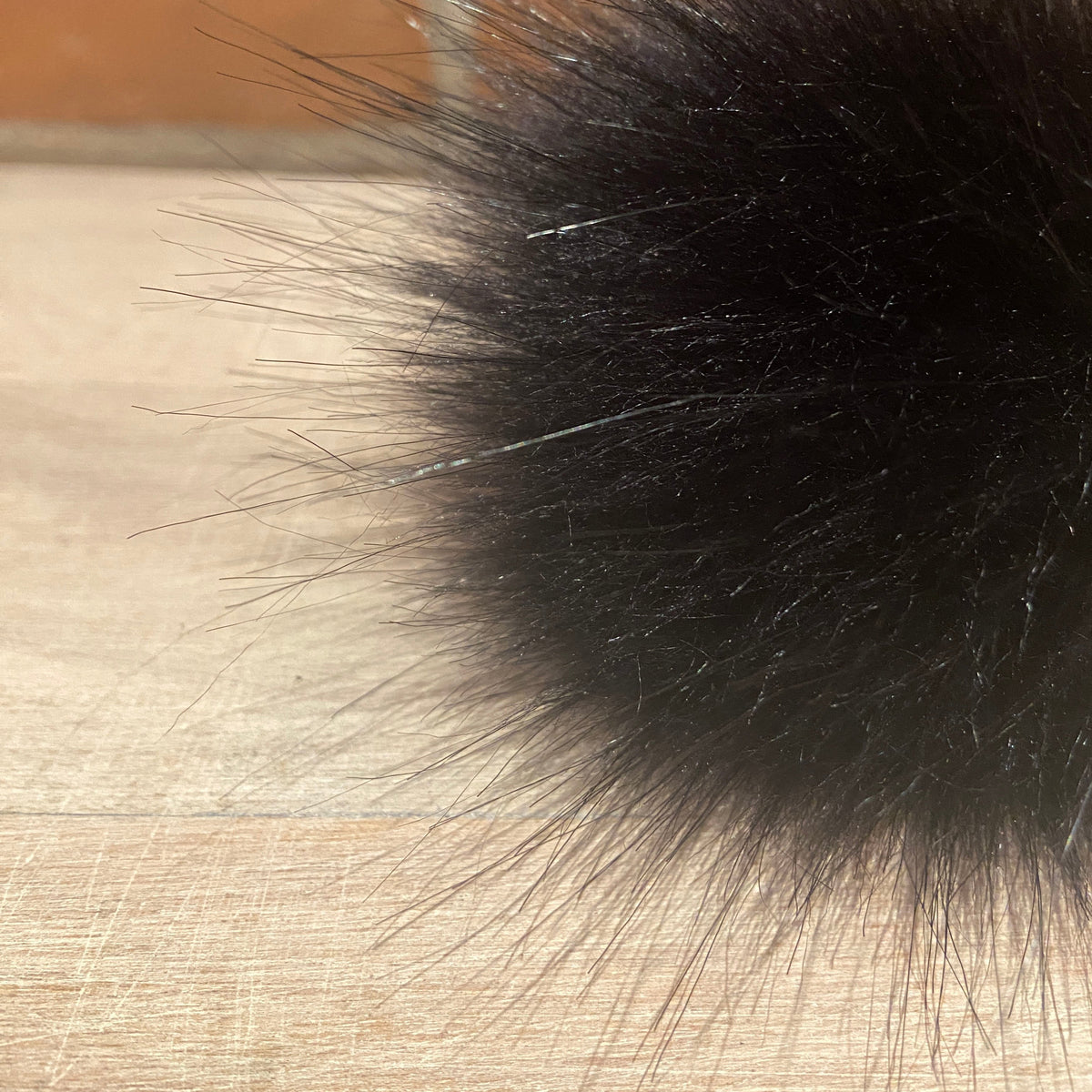 A close-up of a black faux fur pom-pom with individual strands visible, resting on a light wood surface with a blurred brick wall background.