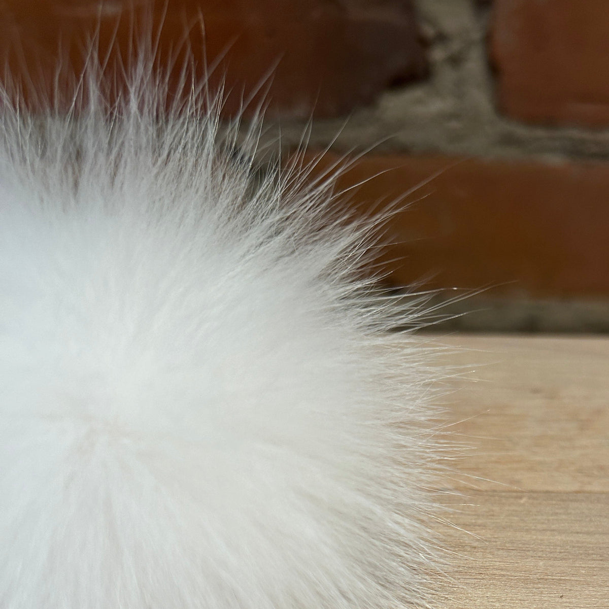 A close-up of a fluffy white fox fur pom-pom resting on a light wood surface with a blurred brick wall background.