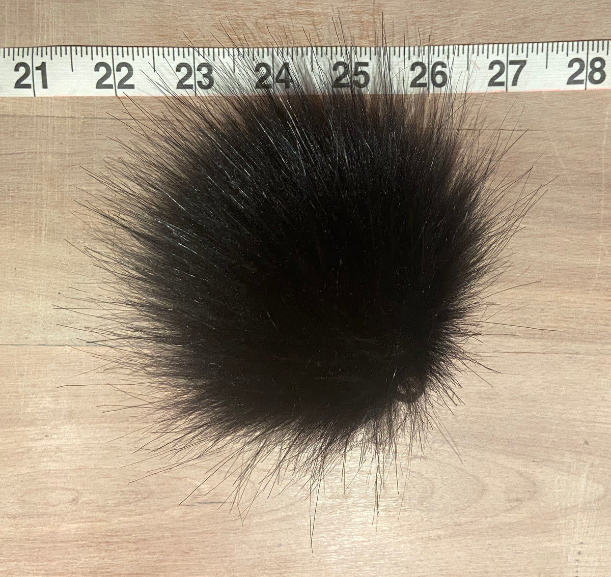 A black faux fur pom-pom ball shown on a light wood surface with a measuring tape ruler at the top for scale.