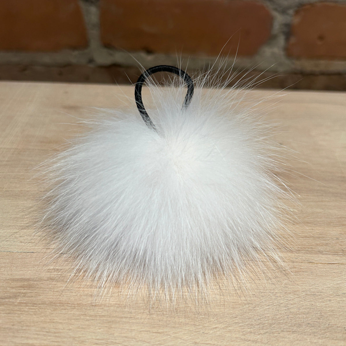 A fluffy white fox fur pom-pom keychain or bag charm with a black elastic loop, resting on a light wood surface with a brick wall background.
