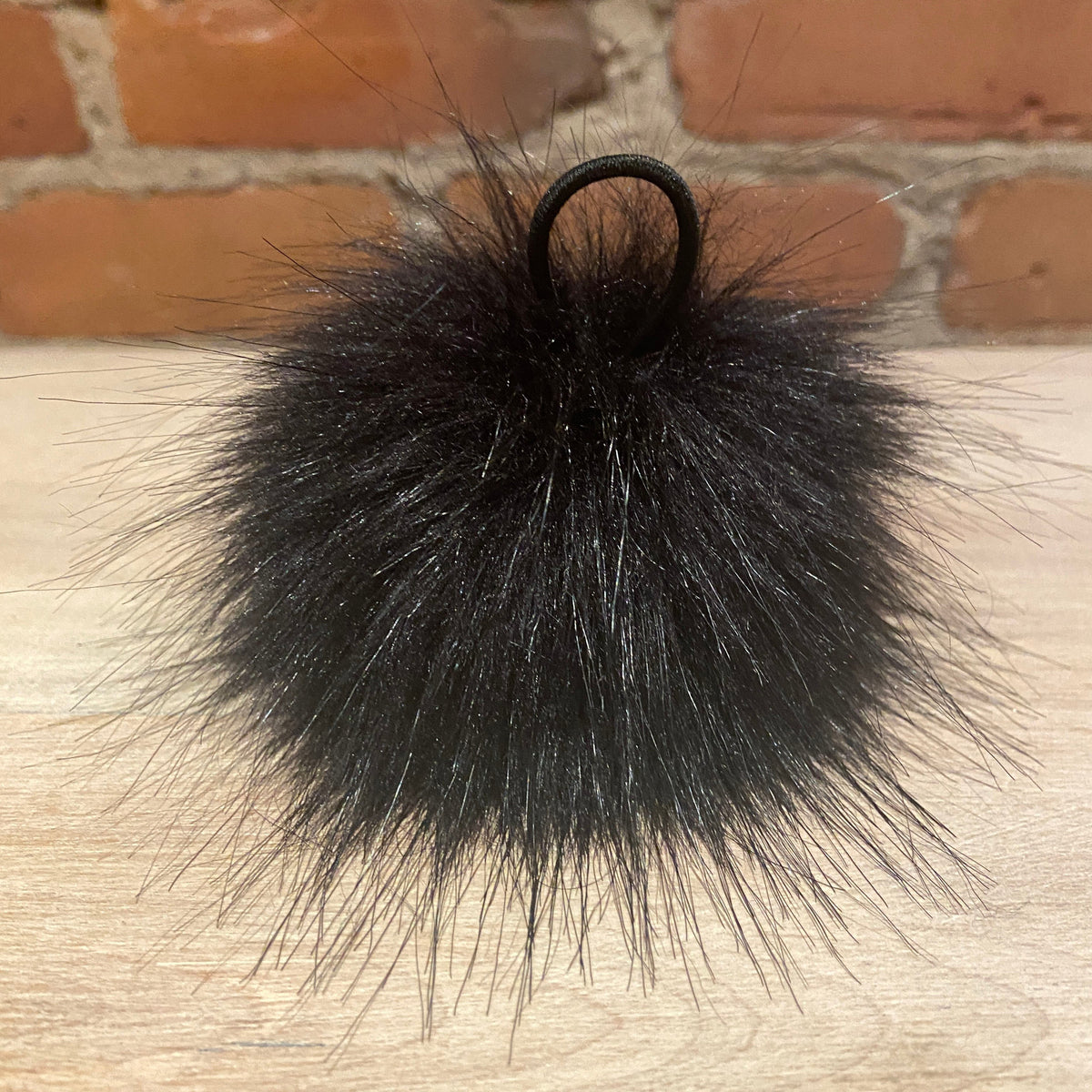 A black fluffy faux fur pom-pom keychain or bag charm with a black elastic loop, resting on a light wood surface with a brick wall background.