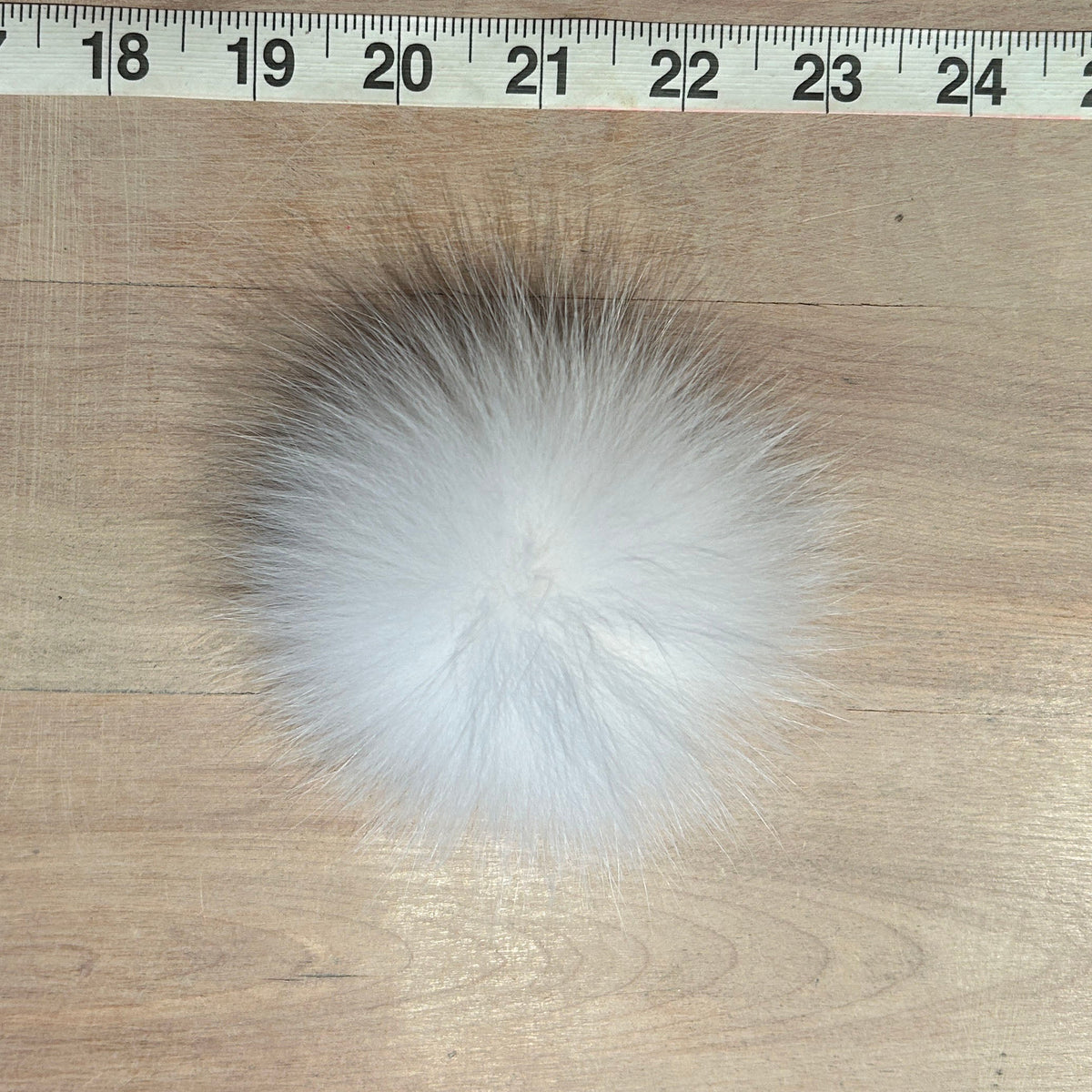 A fluffy white fox fur pom-pom ball shown on a light wood surface with a measuring tape ruler at the top for scale.
