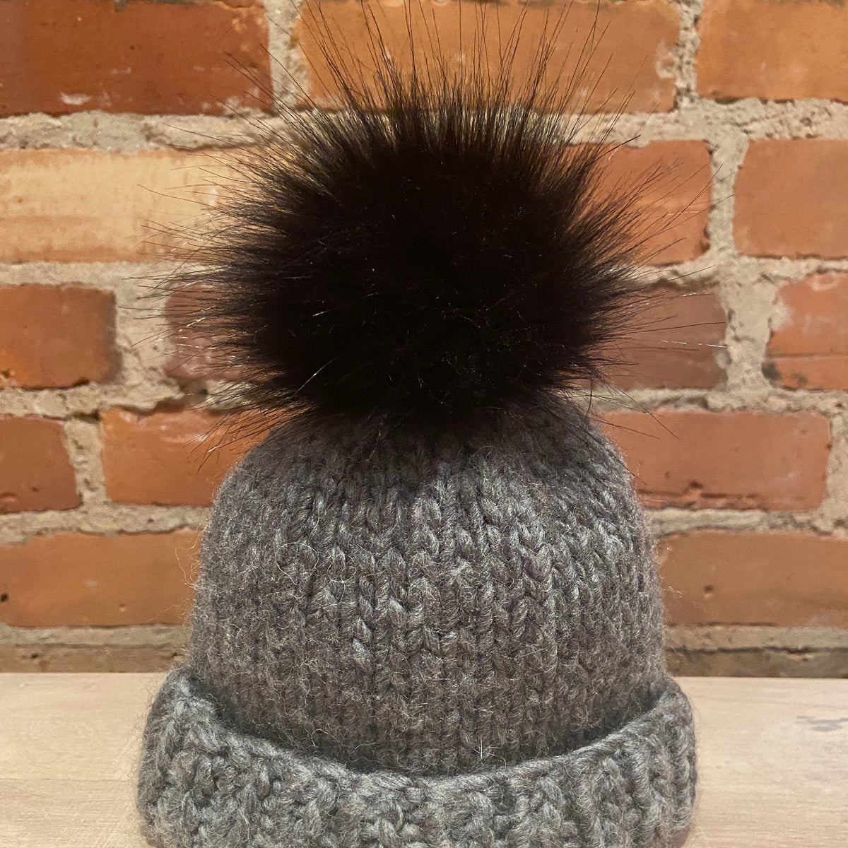 A small gray chunky knit beanie hat with a small black faux fur pom-pom, displayed on a light wooden surface against a red brick wall background.