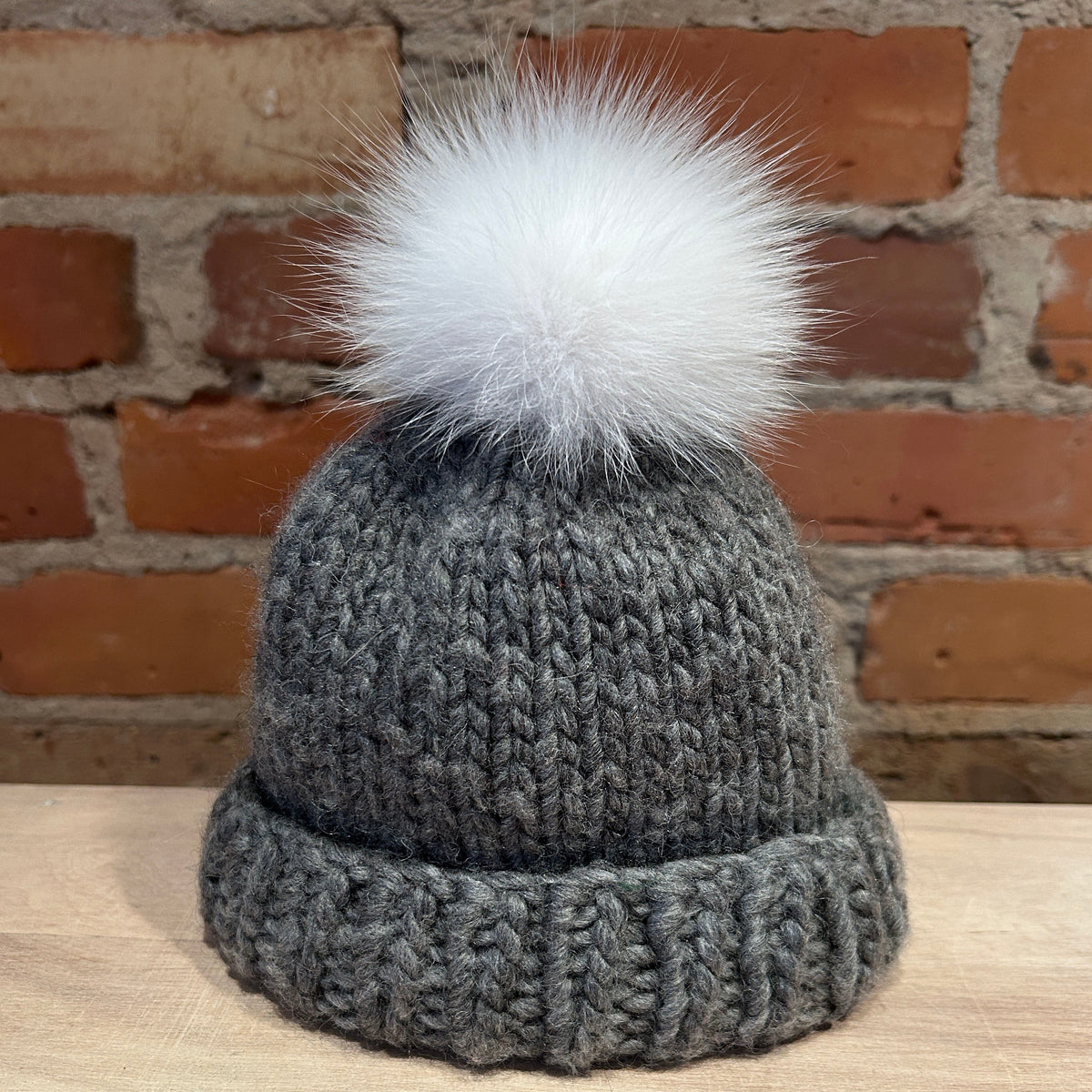 A small gray ribbed knit beanie hat with a small fluffy white pom-pom, displayed on a light wood surface against a rustic red-brown brick wall background.