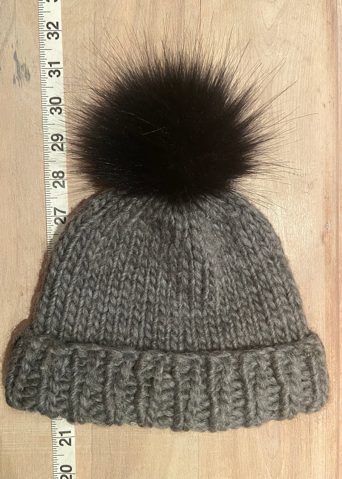 A small gray ribbed knit beanie hat with a small black faux-fur pom-pom, laid flat on a light wood surface next to a measuring tape.
