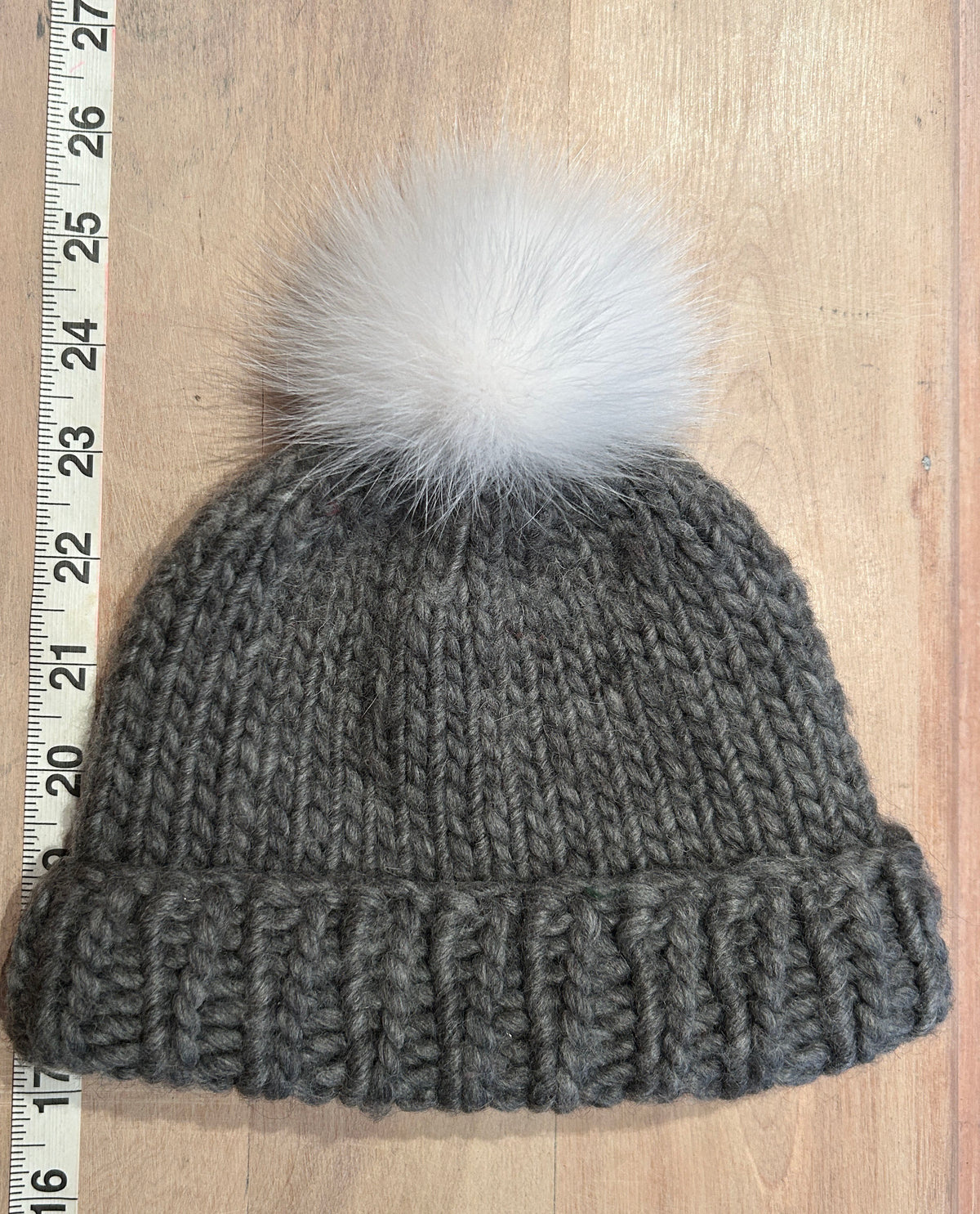 A gray chunky knit beanie with a fluffy white pom-pom, laid flat on a light wood surface next to a measuring ruler.