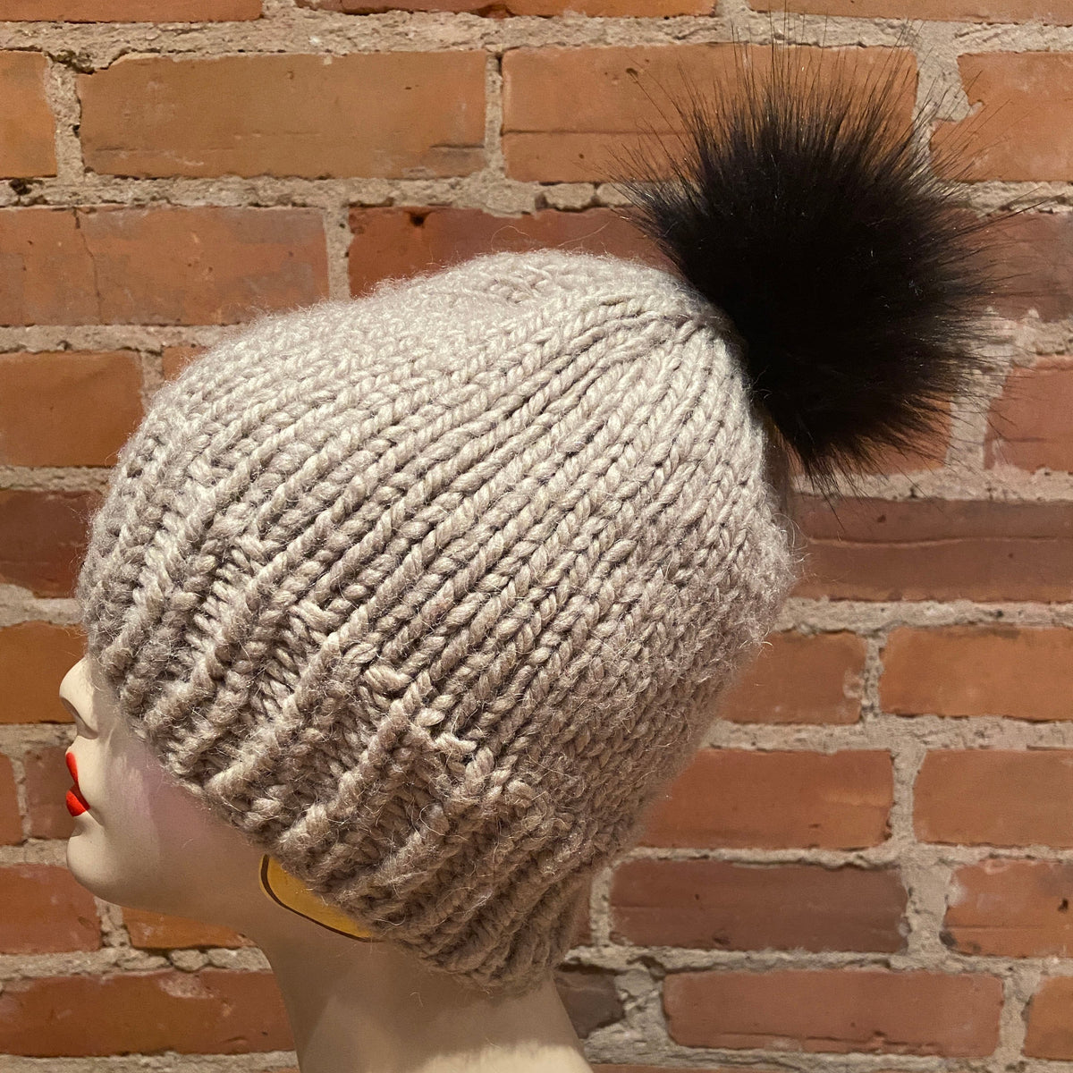A light beige chunky knit beanie with a small black faux fur pom-pom, displayed on a mannequin head against a red brick wall background.