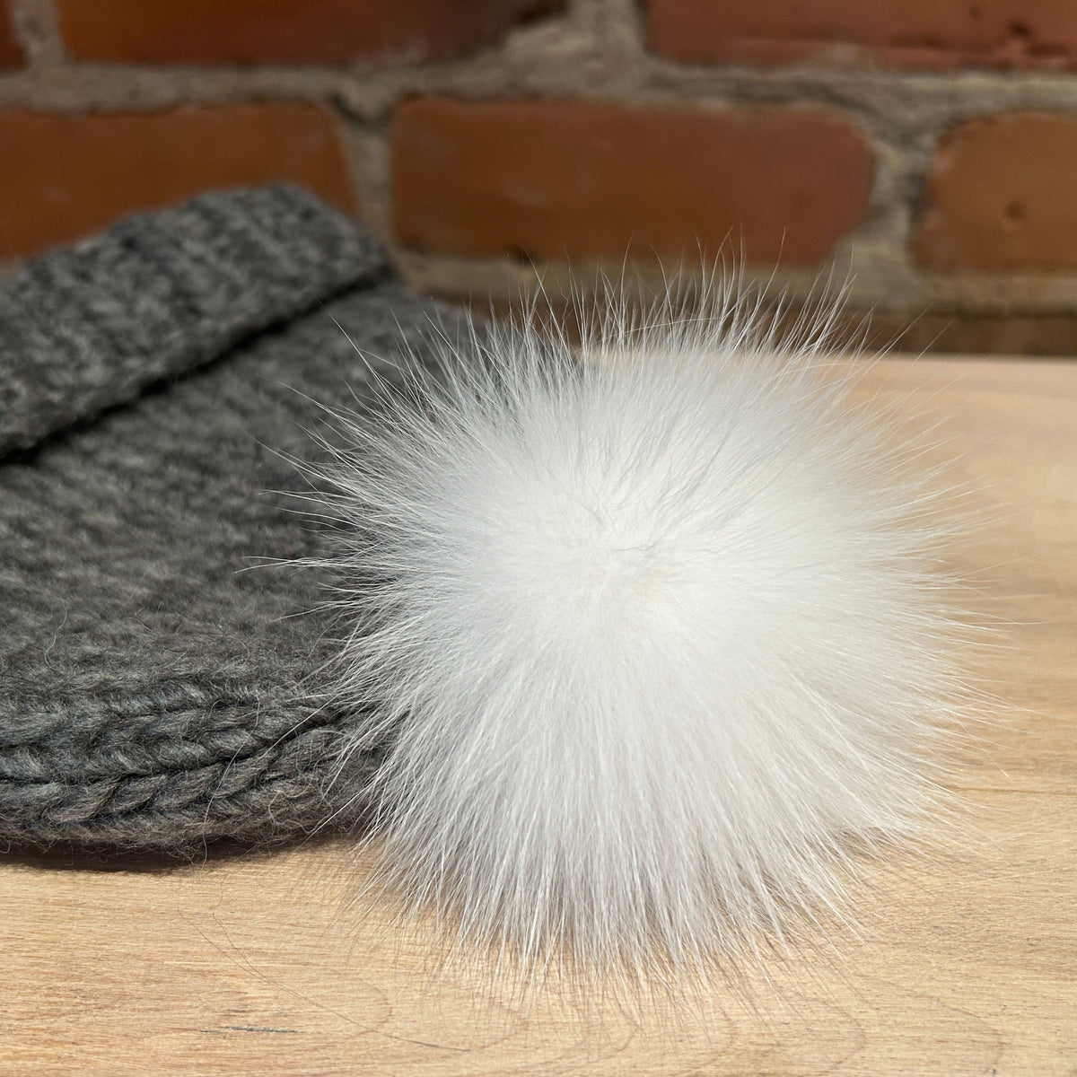 A close-up of a gray knit beanie with a fluffy white fox-fur pom-pom, resting on a light wood surface with a brick wall background.