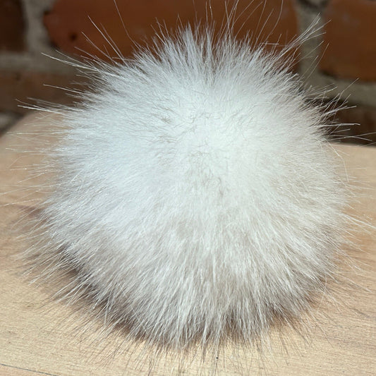 A fluffy white faux fur pom-pom ball resting on a light wood surface with a blurred brick wall background.