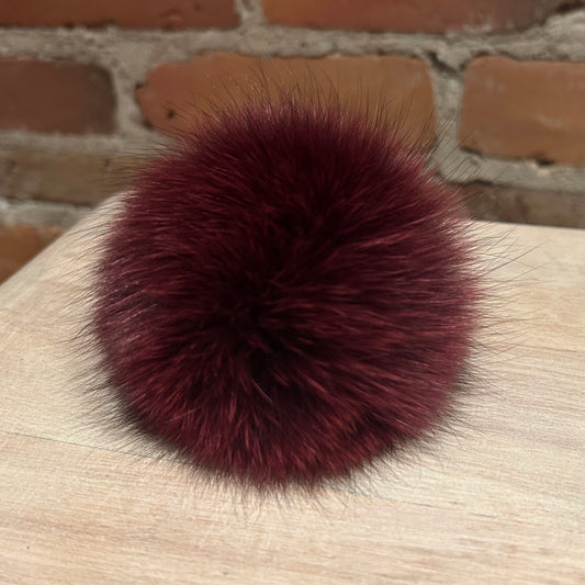 A round, fluffy pom-pom accessory in deep burgundy faux fur, resting on a light wood surface with a brick wall background.