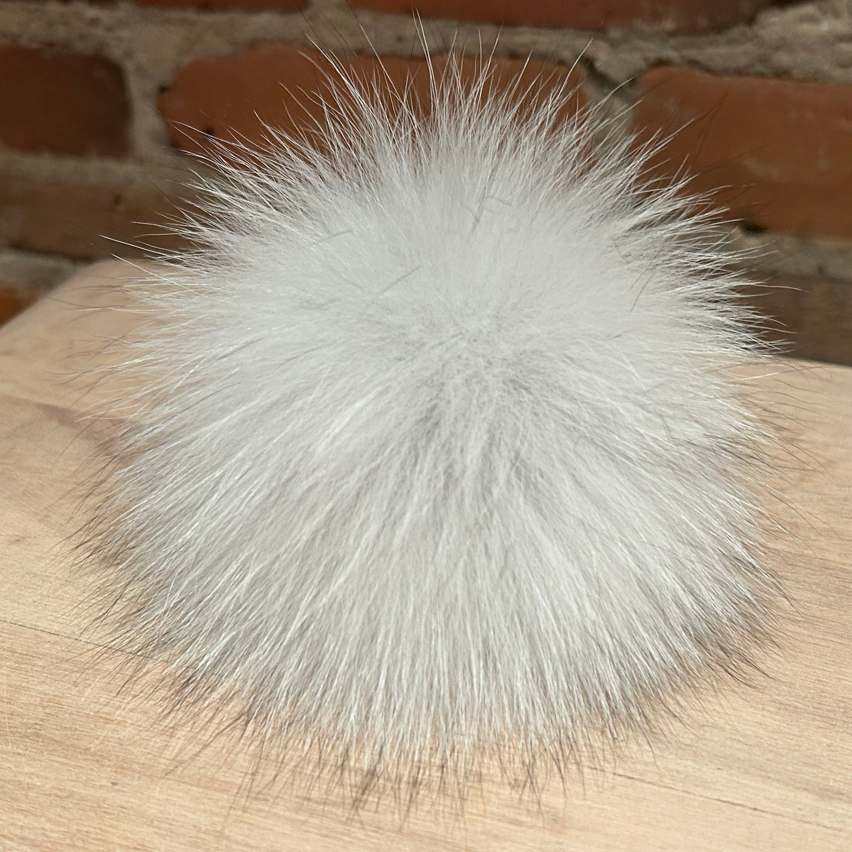 A fluffy white fox fur pom-pom ball resting on a light wood surface with a blurred brick wall background.