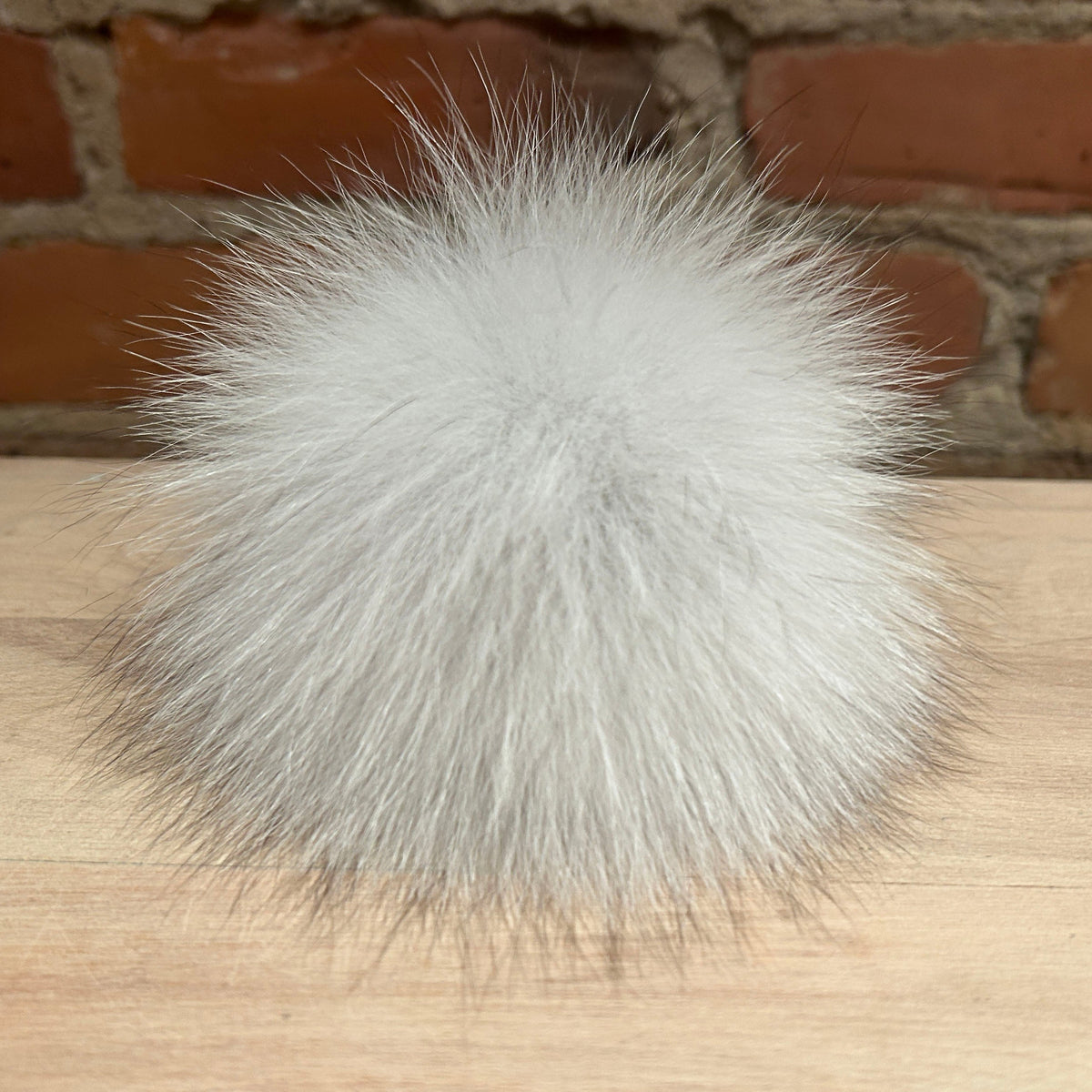 A fluffy white fox fur pom-pom ball resting on a light wood surface with a brick wall background.